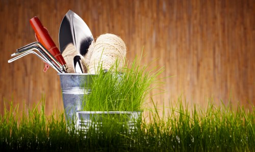 The image features a close-up view of a patch of bright green grass in the foreground, typical of a well-maintained lawn within a garden setting. Behind the grass, there are two metal gardening containers, one slightly larger, holding various gardening tools such as a garden fork, a trowel, and a pair of pruning shears. A ball of twine and a lush, green plant with thin, grass-like leaves are also visible in one of the containers, suggesting preparation for outdoor gardening or maintenance activities. The background consists of a wooden surface, likely a garden fence or shed wall, with warm, natural tones complementing the fresh greenery and indicating a well-kept outdoor space common in residential gardens around Hounslow. The overall scene captures a moment of garden readiness, with tools and plants prepared for trimming, planting, or general exterior maintenance, aligning with gardening services provided by companies like Hedge Trimming Hounslow.