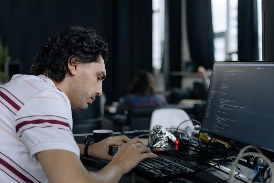 A young man with dark, wavy hair wearing a white shirt with maroon stripes, is seated at a desk in an indoor workspace, focused on his computer screen displaying lines of code. The desk is cluttered w