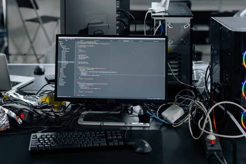 A young man with dark, wavy hair wearing a white shirt with maroon stripes, is seated at a desk in an indoor workspace, focused on his computer screen displaying lines of code. The desk is cluttered w